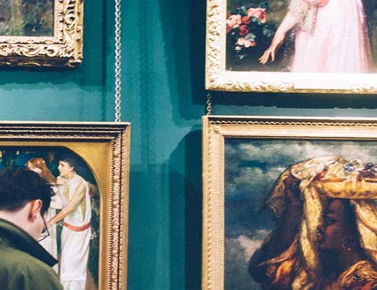 Framed paintings hanging on a teal museum wall, with a visitor partially visible at left