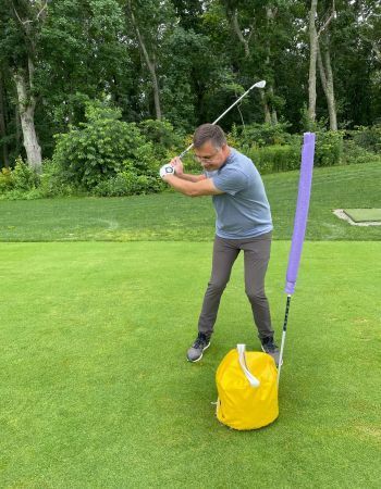 Man swinging golf club on green, purple guide, yellow bag, outdoors.