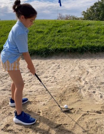 Young golfer in blue shirt and shorts, swings club in a sand trap.