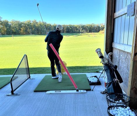 Person swinging golf club at a driving range. Red alignment aid visible. Sunny outdoor setting.