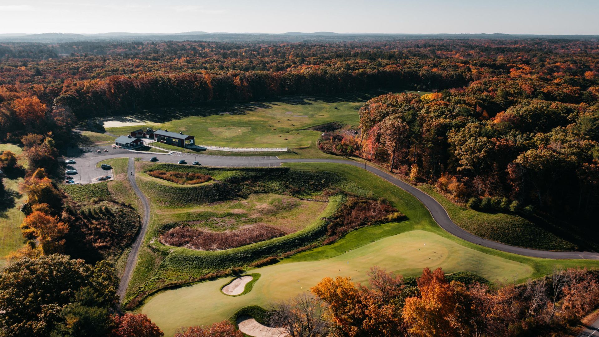 Aerial view of a golf course in autumn; trees with colorful leaves surround green fairways, bunkers, and a building.