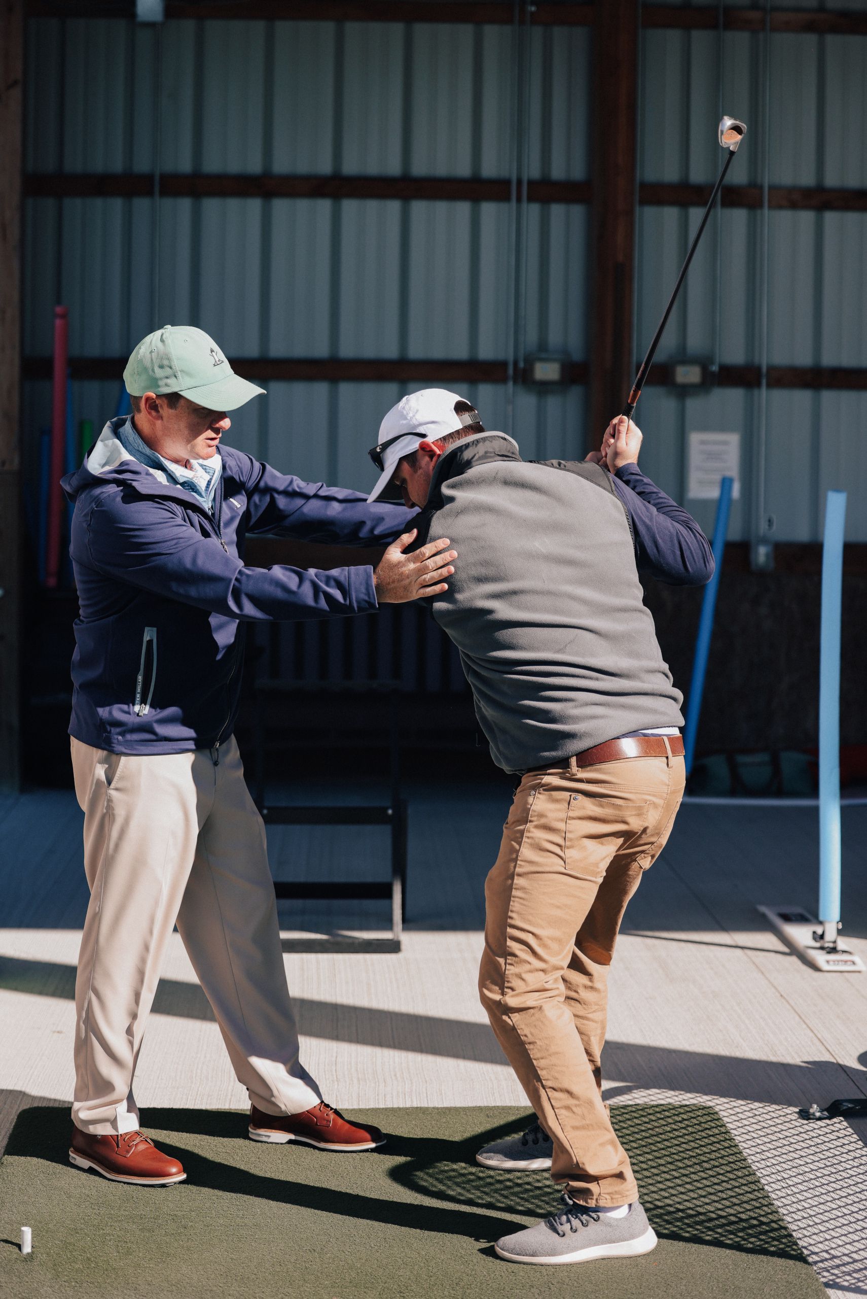 Golf instructor coaching a student's swing, guiding their arms and torso. Indoors, sunny.