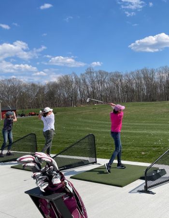 People golfing at a driving range on a sunny day. Green grass, blue sky, and golf equipment in view.