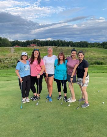 Group of women posing on a golf course green, smiling with clubs, wearing colorful golf attire. Cloudy sky backdrop.