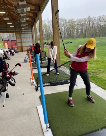 Woman swinging golf club at a driving range, another person in background. Green grass and concrete, covered area.