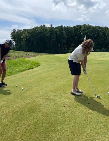 Two people playing golf on a green course under a partly cloudy sky. One swings, the other watches.