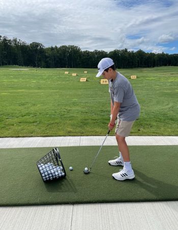 Person in a white hat swings a golf club on a driving range with golf balls.
