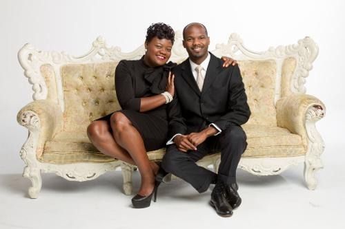 Two people seated on an ornate cream sofa, smiling in a studio setting.
