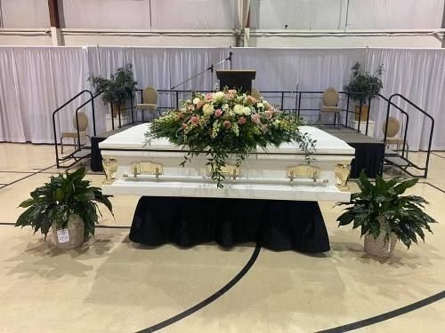 White casket with floral arrangement on a raised stage in a memorial service venue