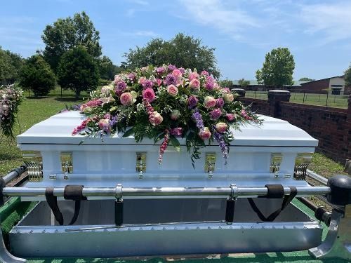 White casket with pink flower spray outdoors beside a lake and fence