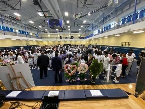 Crowded indoor gymnasium with a stage, floral display, and people gathered for an event