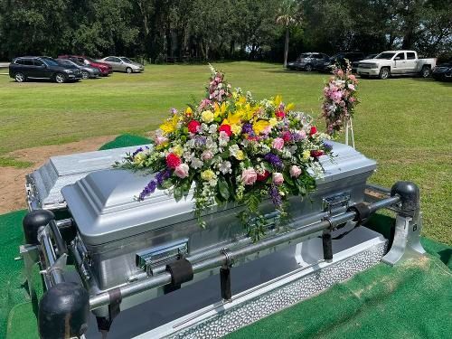 Silver casket with colorful flowers at a graveside memorial, surrounded by green grass and parked cars.