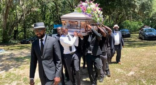 Mourners carrying a flower-covered casket at an outdoor funeral procession, with one man leading ahead.