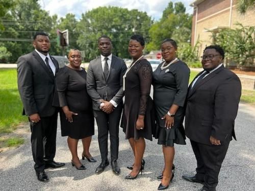 Six people in black formal wear standing together outdoors on a campus lawn.