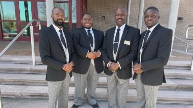 Four men in black blazers and ties stand on steps outside a building, posing together.