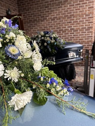Blue and white floral arrangement beside a black casket in front of a brick wall
