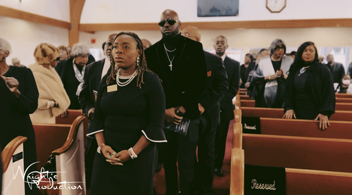 People standing in church pews during a solemn service, with a man in sunglasses at center.