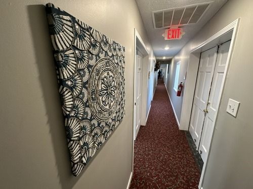Dim hallway with red carpet, white doors, wall art, and an exit sign at the far end.