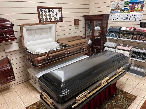 Funeral showroom with an open casket, display cabinets, and stacked caskets on carpeted floor.