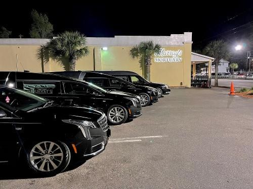 Night parking lot with several black cars lined up beside a lit building and orange cones.