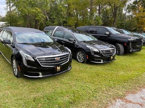 Three black Cadillac sedans parked on grass by trees, front views angled toward the camera.