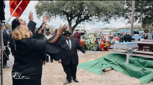 People in black clothing reaching toward a graveside casket covered in green cloth