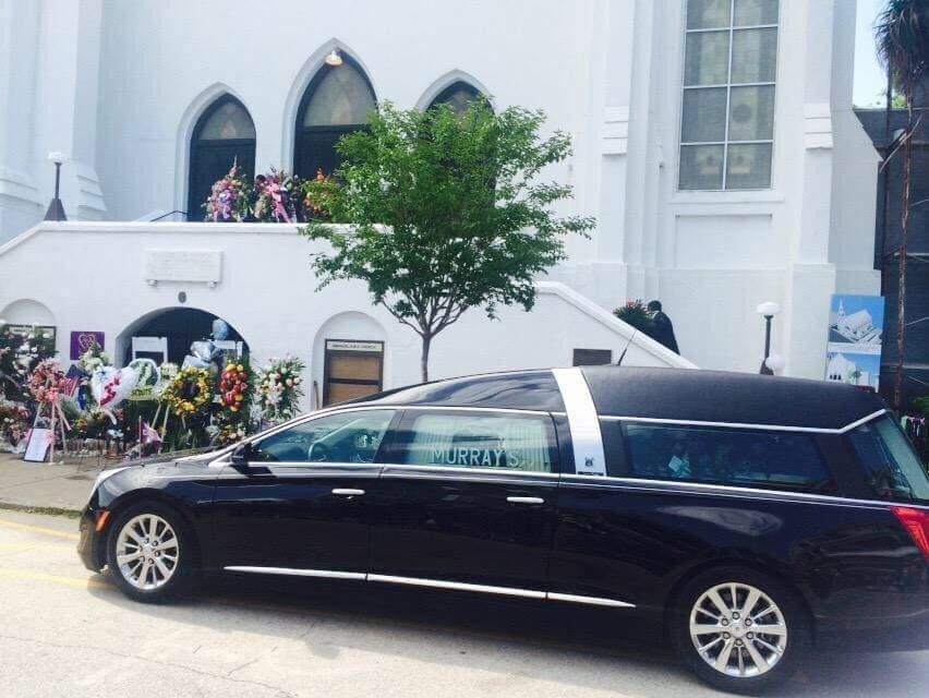 Black hearse parked outside a church with floral tributes and a small crowd nearby