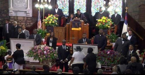 People at a formal memorial service inside a church, with floral arrangements and speakers onstage.