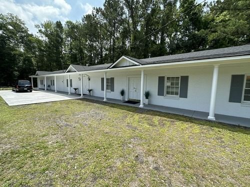 White single-story house with a long porch, a driveway, and a grassy yard beside trees