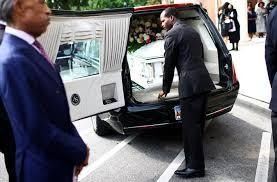 A black hearse with an open rear door as a man loads a white casket, with people standing nearby.