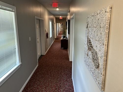Narrow hotel hallway with red carpet, white walls, windows, and overhead exit signs.