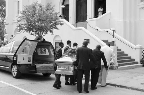 Pallbearers carry a casket from a hearse toward a church entrance on a staircase.