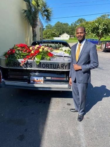Man in gray suit standing beside flower-covered hearse with “MURPHY’S MORTUARY” sign on sunny street