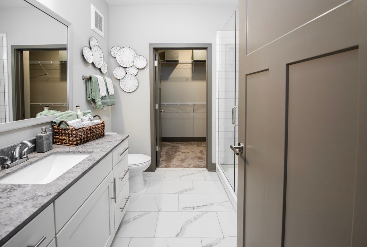 Modern apartment bathroom with white cabinets, marble countertop, sink, toilet, glass shower, and open closet.