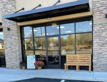 Storefront with large glass windows, stone facade, potted plants, and a wooden bench outside.