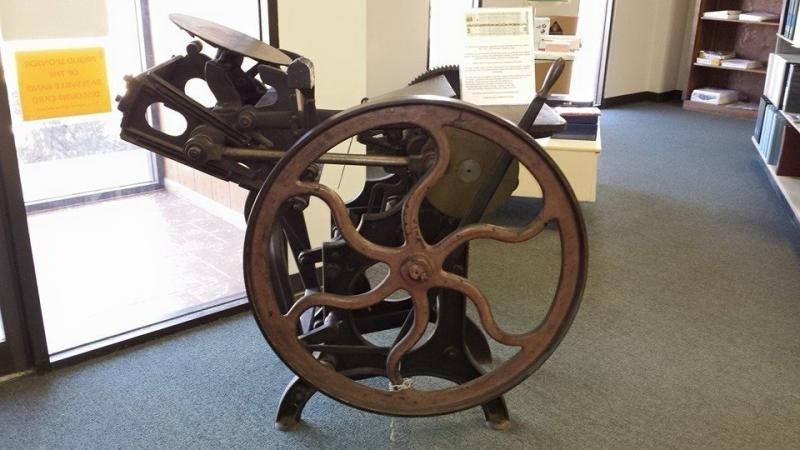Rusty antique machine with a large wheel displayed indoors on a carpeted floor.