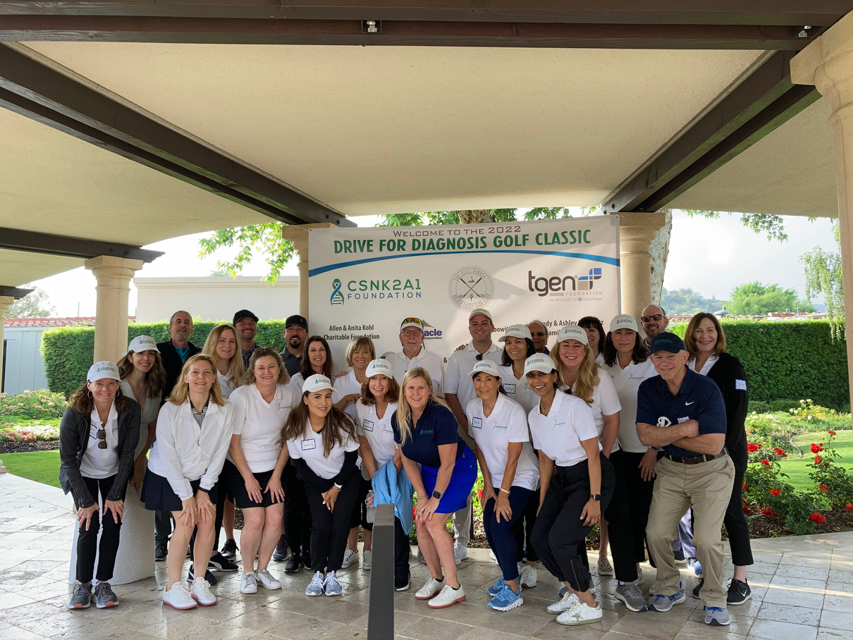 Group of people pose for a photo under an awning at a golf event; banner says
