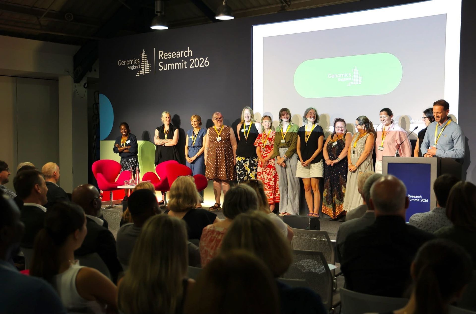 Awards ceremony on stage with presenters lined up, audience seated in foreground, and a projected screen behind them