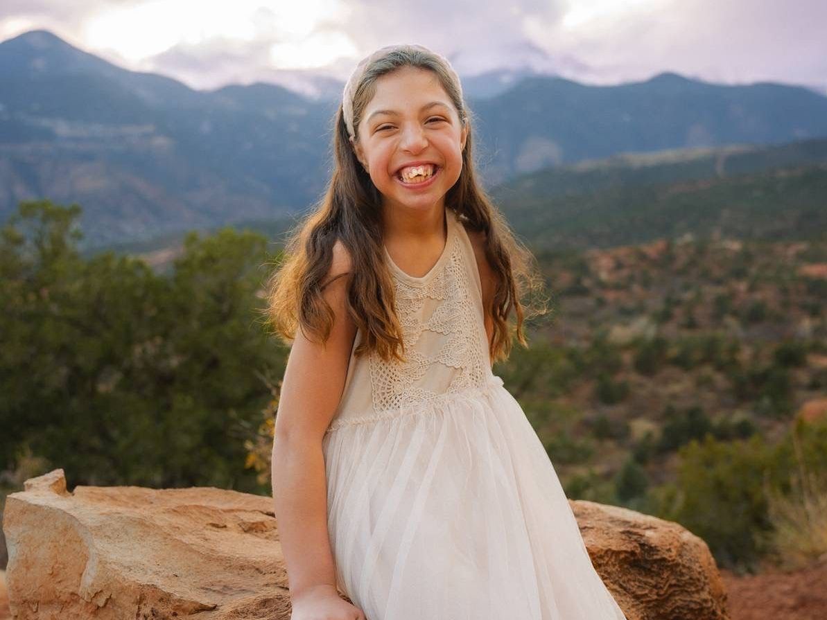 A little girl in a white dress is sitting on a rock with mountains in the background.