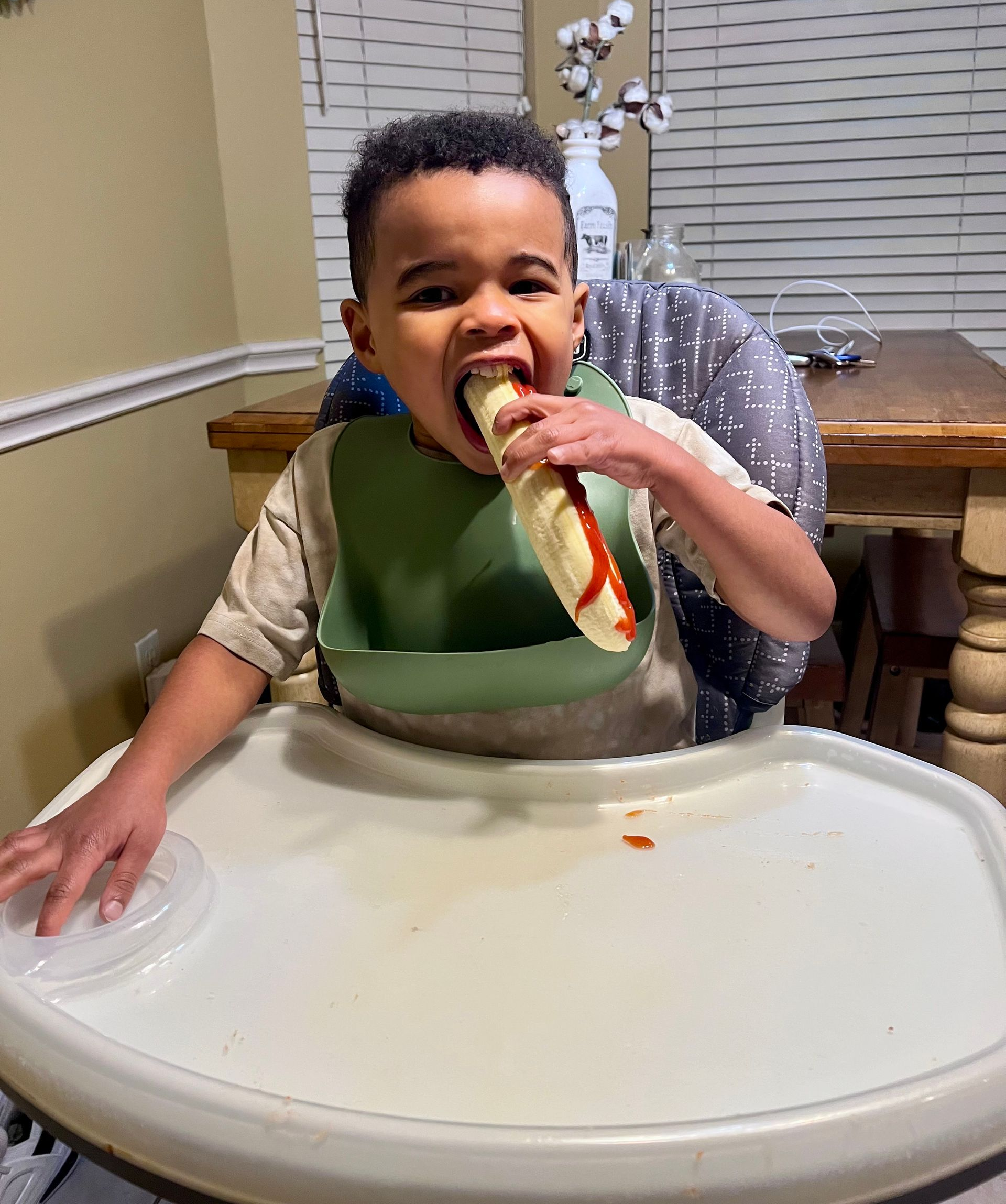 Child in high chair eating a hot dog with ketchup, wearing a green bib.