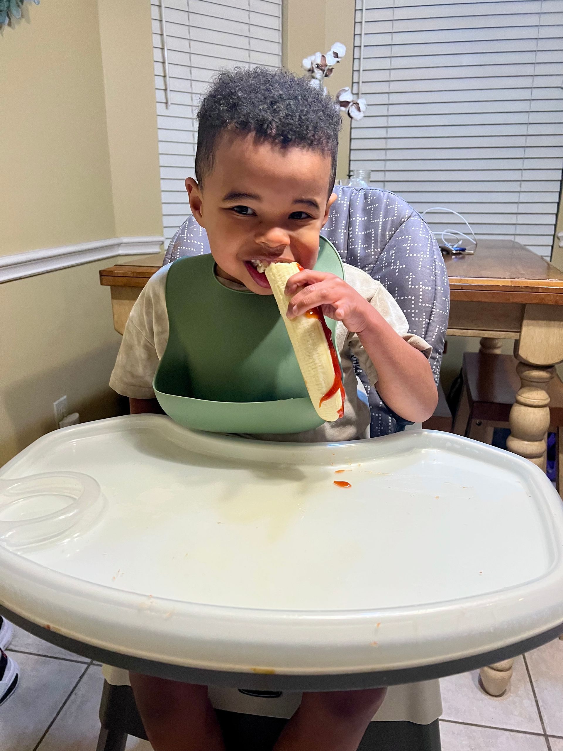 Child in a high chair eating a wrap with ketchup, smiling. Green bib, white tray.