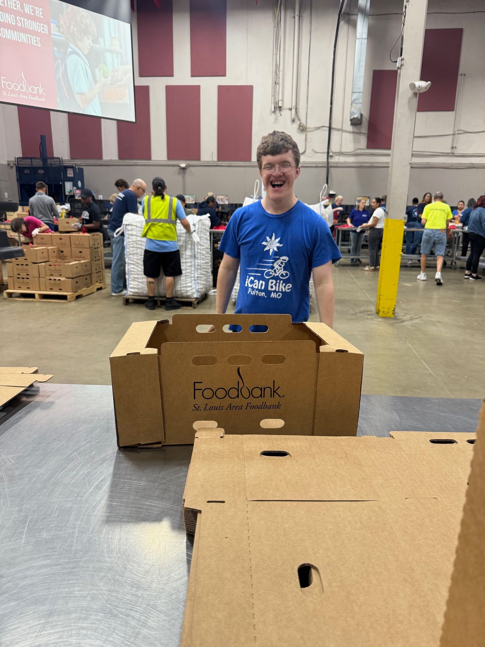 A person smiling at the camera, standing behind a cardboard box at an event.