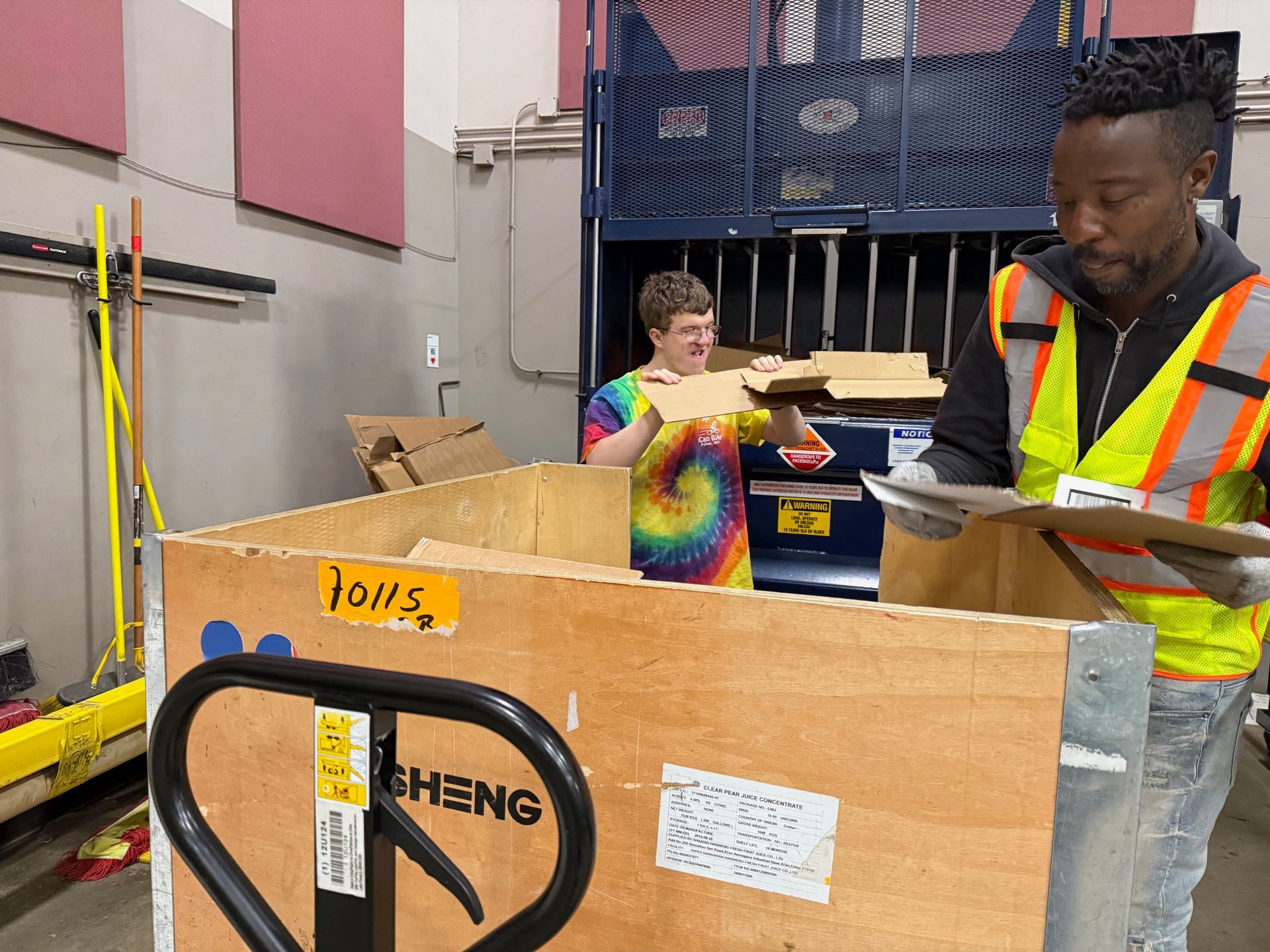 Two people sorting materials in a warehouse. One wears a reflective vest, the other a tie-dye shirt.