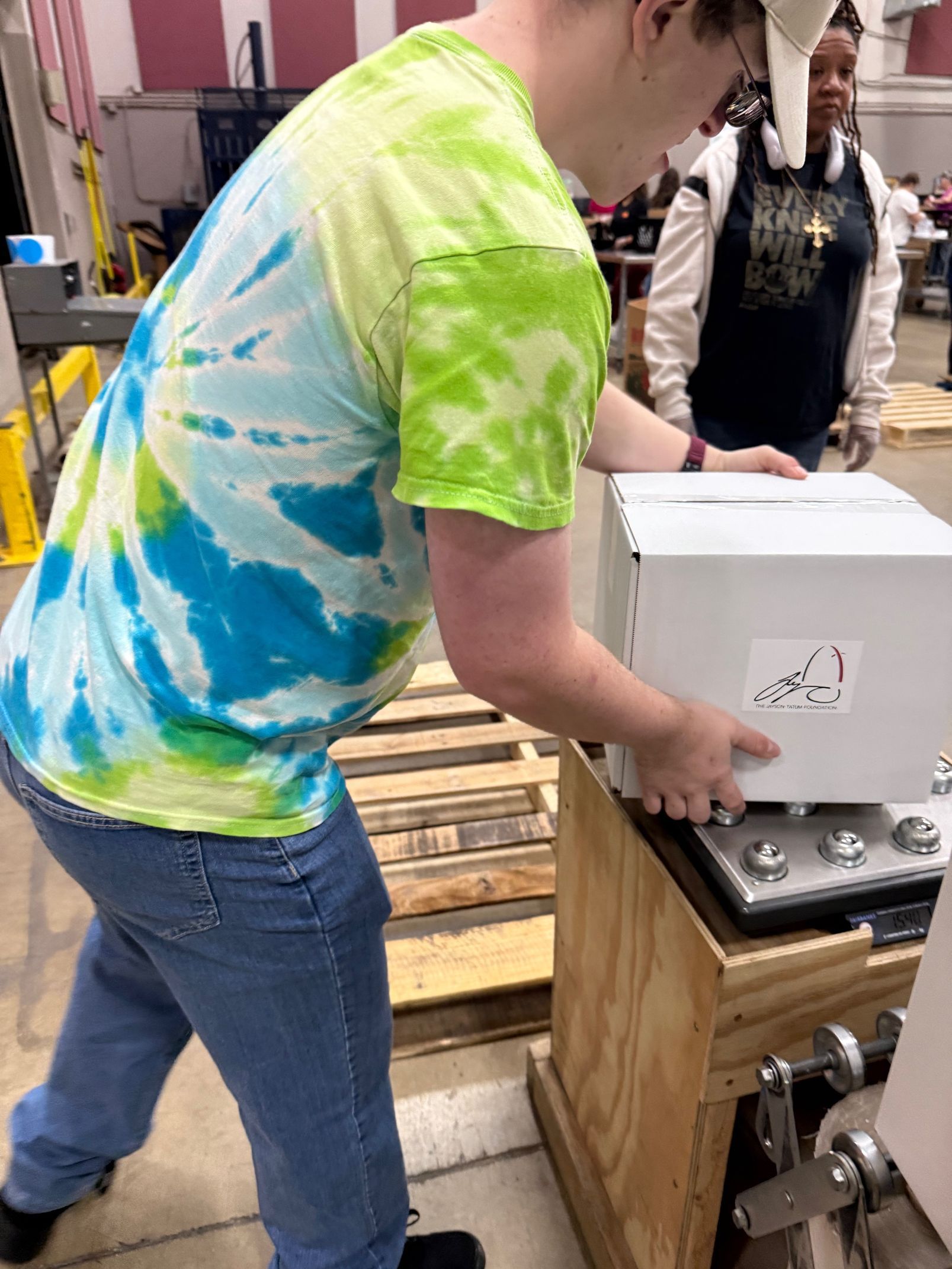 Person in tie-dye shirt lifts a box onto a machine. Another person watches in a room.