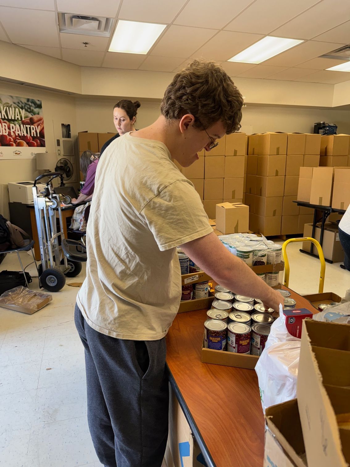 A person sorting canned goods on a table; another person and boxes are in the background.
