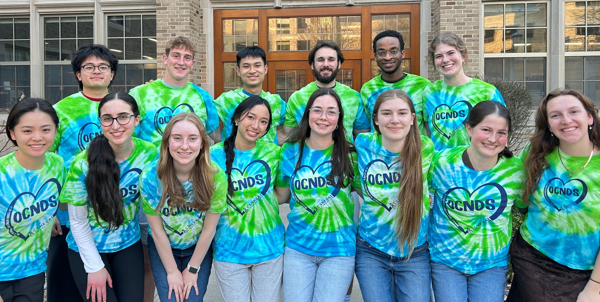 Group standing outside a building, wearing matching blue-green tie-dye shirts and smiling at the camera.