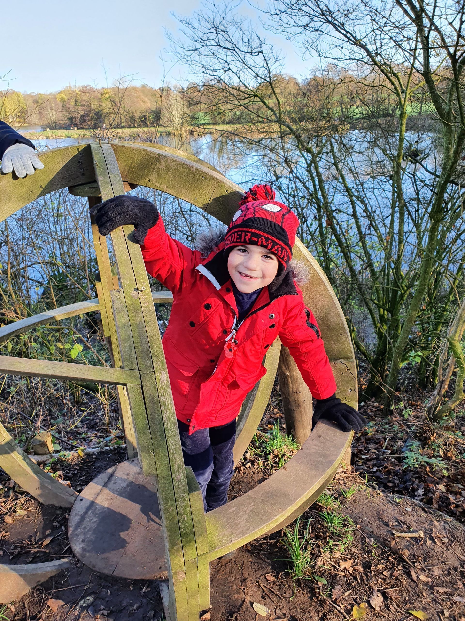 A young boy in a red jacket and hat is sitting on a wooden wheel.