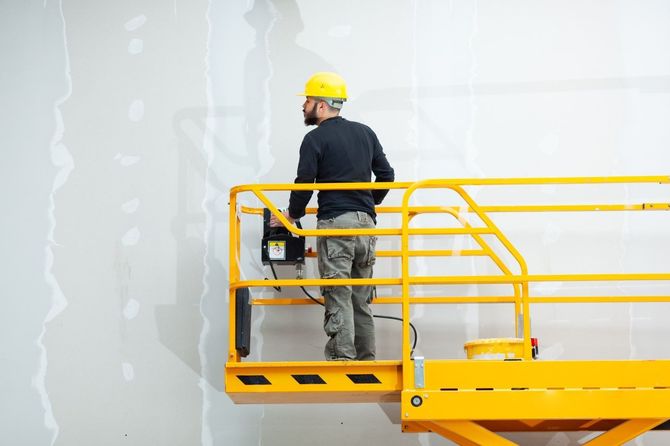 Worker in a yellow hard hat stands on a yellow lift platform painting a wall.