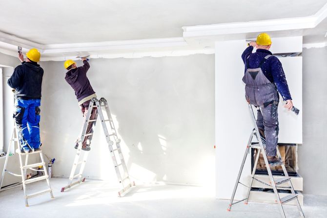 Three workers in hard hats paint a white room, standing on ladders.