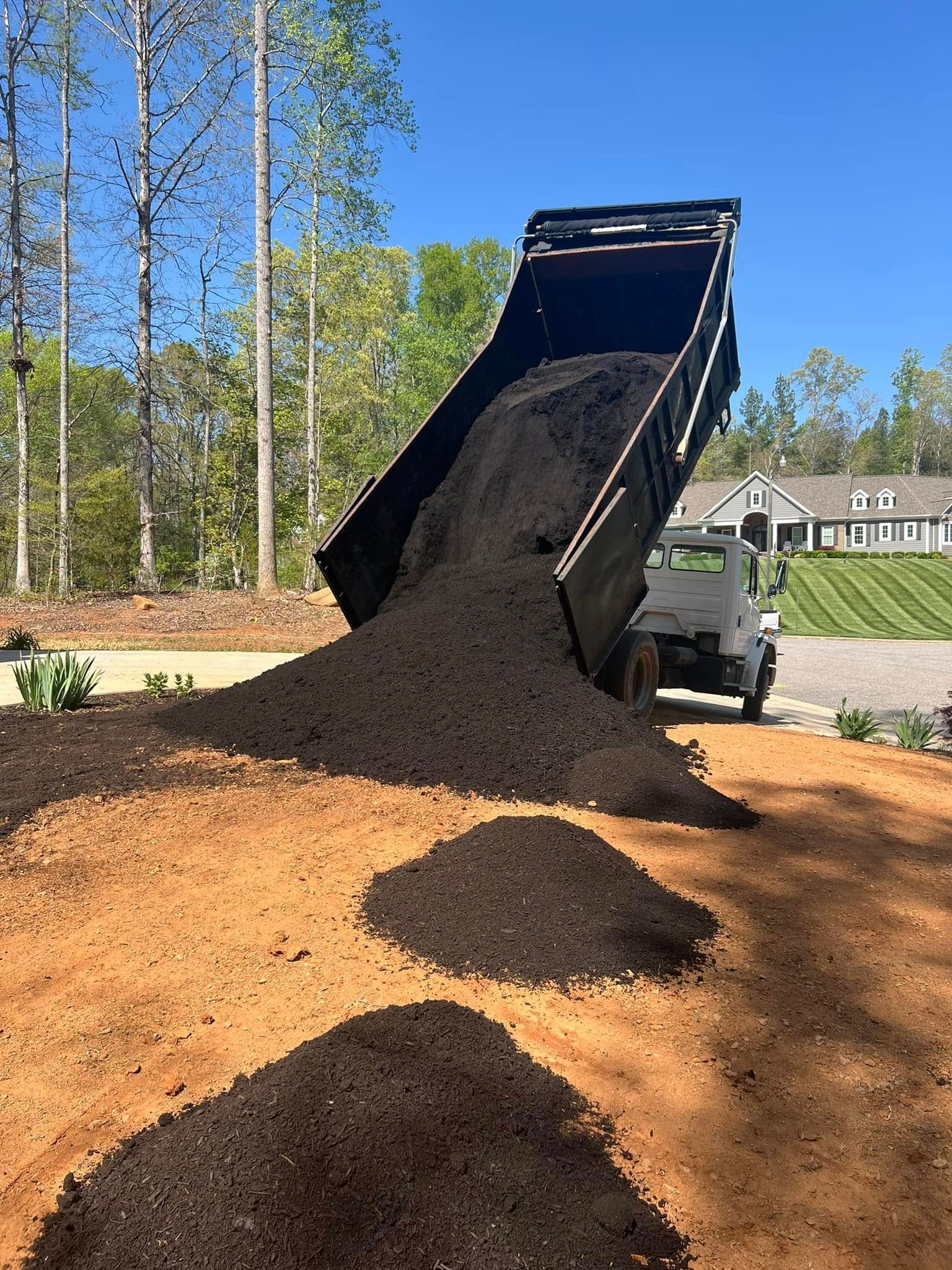 Dump truck unloading mulch on a red dirt area; sunny day, trees in background.
